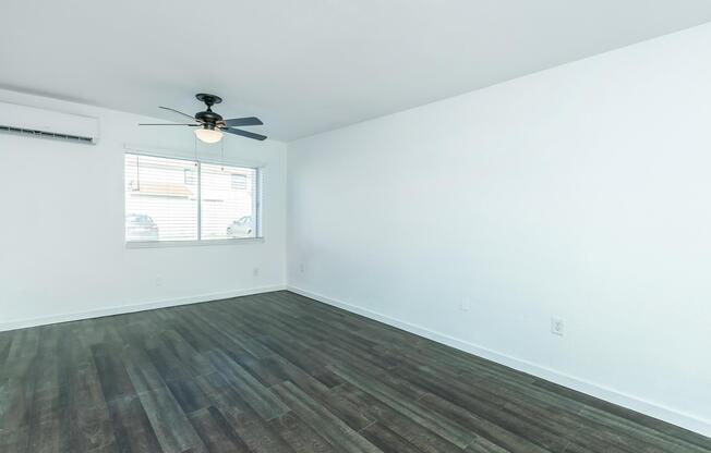 A bright and airy room featuring light-colored walls and dark wood-like flooring. It has a ceiling fan and a window with light blinds, providing natural light. The overall atmosphere is spacious and modern, suitable for various interior designs.
