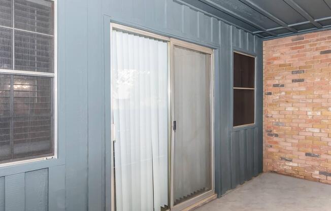 A view of a patio area featuring a sliding glass door with vertical blinds, alongside two windowsâone with a grid patternâand a wall with a brick finish. The exterior is painted in a muted blue color, creating a contrasting backdrop against the brick. The scene is well-lit, suggesting a daytime setting.