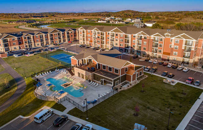 An aerial community view here at The Park featuring modern apartment buildings, a resort-style pool with bright green lounge chairs, tennis court, clubhouse, and landscaped grounds, creating a vibrant, welcoming neighborhood setting.