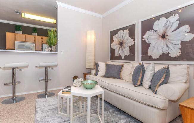 A bright and modern living room featuring a white sofa with decorative pillows, a small white coffee table, and a rug on the carpeted floor. Two bar stools are positioned at a kitchen counter in the background. Large floral artwork hangs on the walls, with greenery in the corner adding a fresh touch.