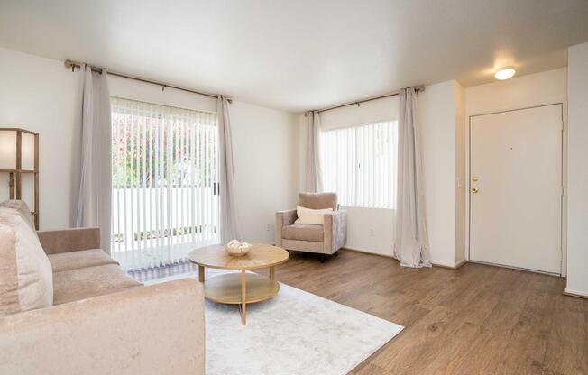 A bright, minimalist living room featuring a light beige sofa and an armchair, a round wooden coffee table, and a light-colored area rug. Two large windows with vertical blinds let in natural light, and soft curtains frame the windows. The entrance door is visible on the right.