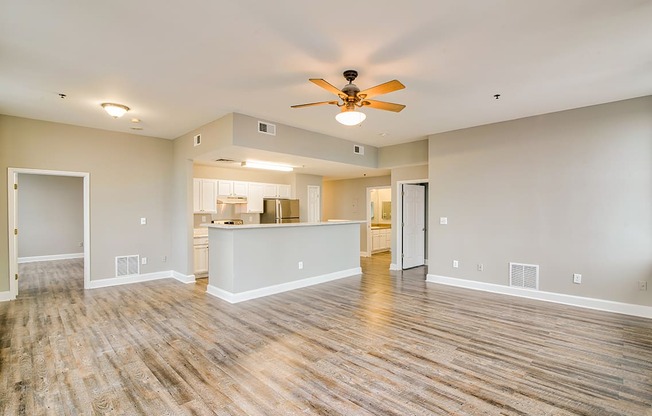 an empty living room with a ceiling fan and a kitchen in the background