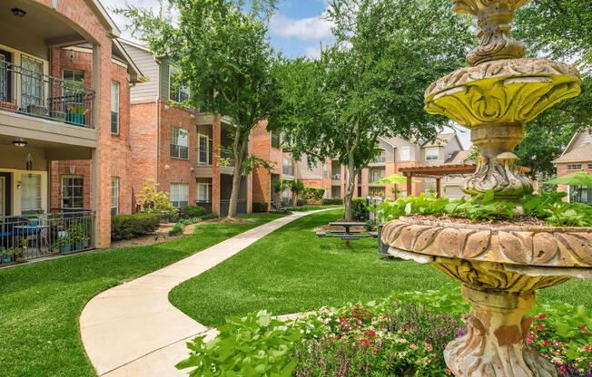 A landscaped courtyard at an apartment complex, featuring a stone fountain surrounded by greenery. The pathway winds through lush grass and flower beds, with brick buildings and trees visible in the background under a blue sky.