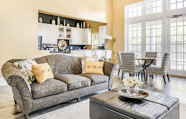 Clubhouse interior with gray couch, ottoman, accent rug and clubhouse kitchen in background with bar top and dining table with chairs