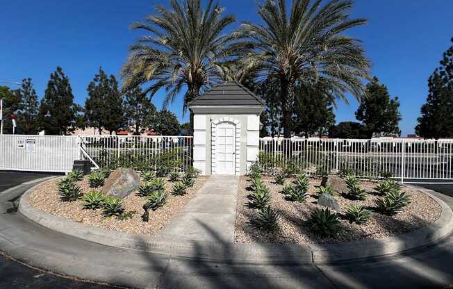 A small white building with a black roof is surrounded by a white fence and palm trees.