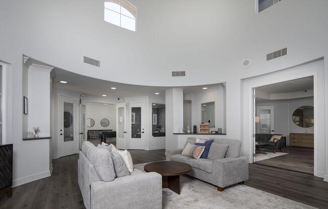 Modern, open-concept living space featuring a circular design. Light-colored walls and hardwood flooring create a bright, airy atmosphere. A cozy gray sofa and circular coffee table are placed in the foreground, with doorways leading to additional rooms and mirrors on the walls reflecting light.