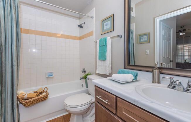 A clean bathroom featuring a white tiled shower with a light brown stripe, a bathtub with a showerhead, a toilet, and a wooden vanity with a sink. Towels are neatly hung, and a woven basket contains rolled towels. The mirror reflects the room, providing a cozy and well-organized appearance.