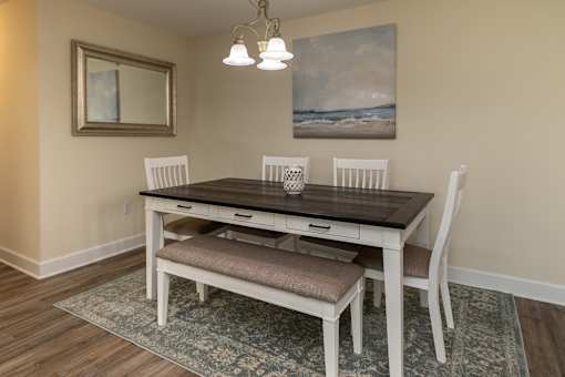 A dining table with four chairs and a bench seat is in a room with a painting on the wall.