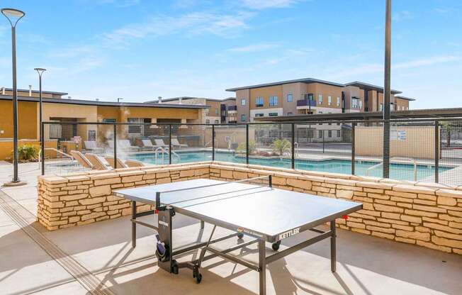 A ping pong table is in the middle of a patio with a pool and a building in the background.