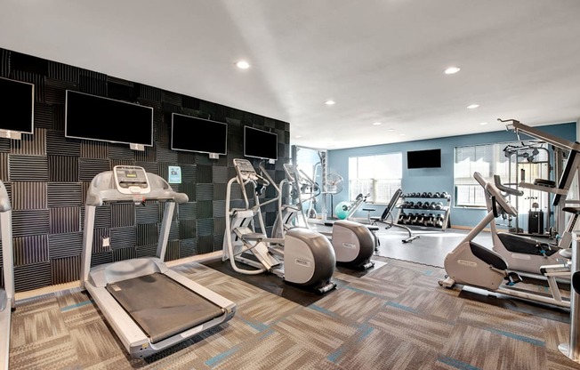 A spacious fitness center here at Vue at Pinnacle Park with treadmills, ellipticals, strength machines, free weights, wall-mounted TVs, and large windows providing bright light for an energizing workout environment.