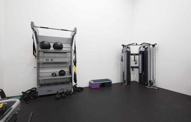 A modern gym space featuring a weight training machine on the right, a storage rack with exercise balls and accessories in the center, and colorful workout mats on the floor. The walls are white, and the floor has a dark rubber surface, creating a clean and functional fitness environment.