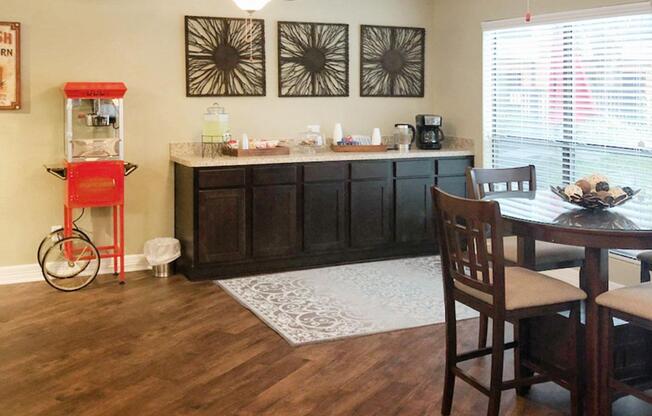 A cozy kitchen or dining area featuring a dark wooden cabinet with a countertop, a popcorn machine in red, a coffee maker, and several cups. There is a small round dining table with four brown chairs, and a decorative rug on the wooden floor. Three decorative wall art pieces are displayed above the countertop, and there are large windows letting in natural light.