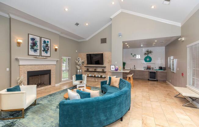A modern lounge area featuring a cozy seating arrangement with teal and white chairs, a fireplace, and a wall-mounted TV. A stylish bar area with a counter, decorative elements, and natural light filters in through large windows, creating an inviting atmosphere.