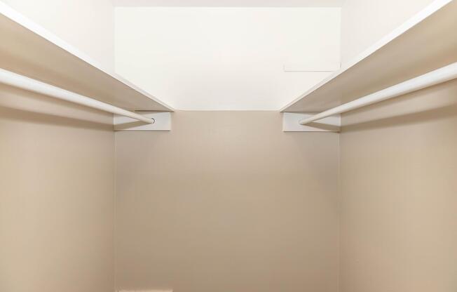 A view inside an empty closet, featuring two white horizontal rods for hanging clothes installed on opposite walls. The walls are painted a neutral beige color, and the ceiling is white. The space appears clean and organized, with no items stored inside.