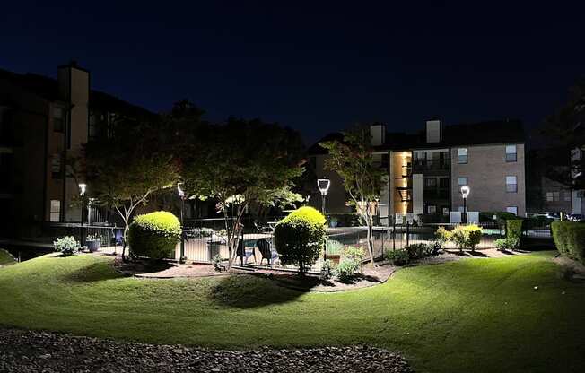 A well-lit park area with a grassy field, trees, and a building in the background.