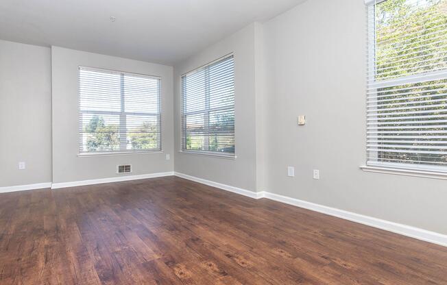 Bright, empty living room with large windows featuring white blinds. Light gray walls and hardwood flooring create a modern and spacious atmosphere. Sunlight streams in, highlighting the opened space, perfect for personal decoration and furniture arrangement.