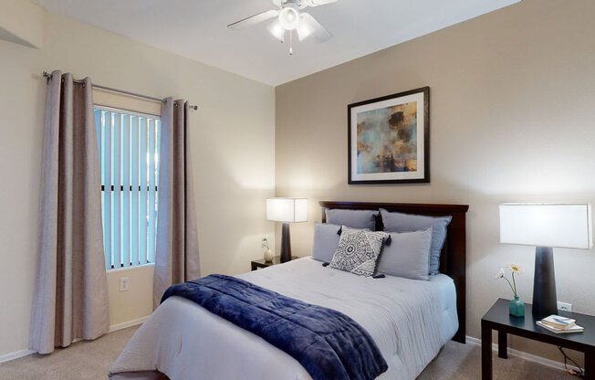 A cozy and tranquil bedroom here at Pala Mesa featuring a plush bed with layered neutral bedding and a soft accent throw, framed by matching bedside tables with modern lamps. A large window with vertical blinds and curtains allows natural light to filter in, while plush carpeting and calming wall tones create a peaceful, comfortable retreat.