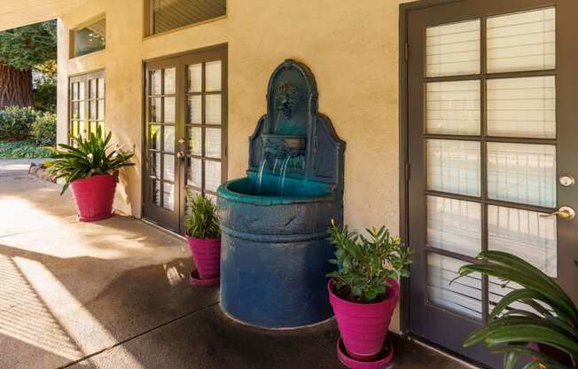 A blue fountain is in the middle of a concrete area with potted plants on each side.