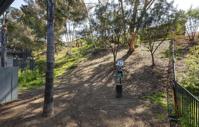 A wooded area with trees and shrubs, featuring a sloped hill. In the foreground, a black post with a sign and a green dispensing unit for dog waste bags. Fencing lines the right side, and a building is partially visible in the background among the trees. The scene is sunny and inviting, typical of a park or natural space.