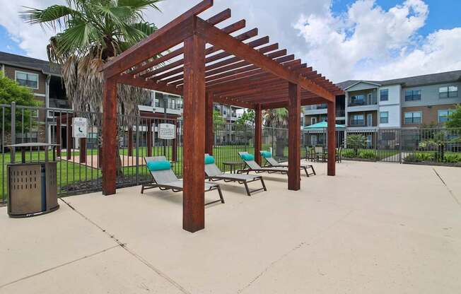 A wooden pergola with blue chairs and a trash can in front of apartment buildings.