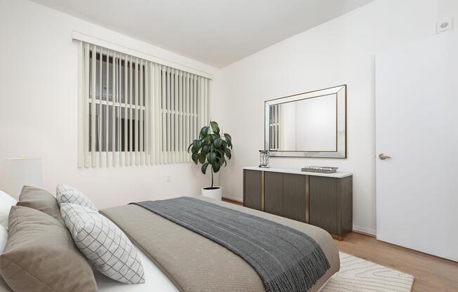 A modern bedroom featuring a neatly made bed with gray and white bedding, two pillows, and a cozy throw. The room has light-colored walls, large windows with vertical blinds, and a stylish mirror above a dark wooden sideboard. A potted plant adds a touch of greenery, enhancing the contemporary ambiance.
