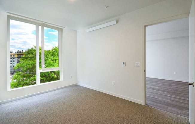A room with a carpet floor, a window with a view of trees and buildings outside, and a closed door. at Ravello Apartments, Redmond