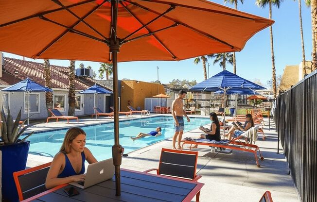 A woman is working on her laptop under an orange umbrella at a poolside table.