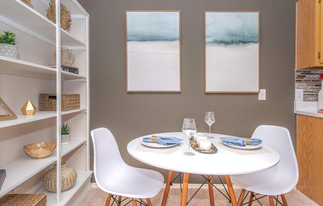 A cozy dining area featuring a round white table set for two with blue napkins and wine glasses. Two simple framed paintings adorn the wall above the table. A white bookshelf is to the left, displaying decorative items and plants, while a kitchen area is visible in the background. The walls are painted in a muted gray tone.