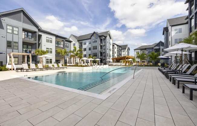 A large swimming pool surrounded by lounge chairs and umbrellas in front of apartment buildings.