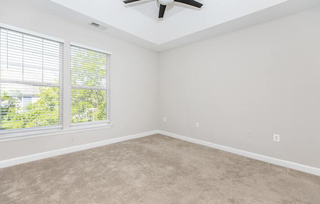 A spacious, empty room with light-colored walls and soft beige carpet. It features a ceiling fan and large windows with white blinds, allowing natural light to fill the space. The room is well-lit and appears ready for decoration or furnishing.