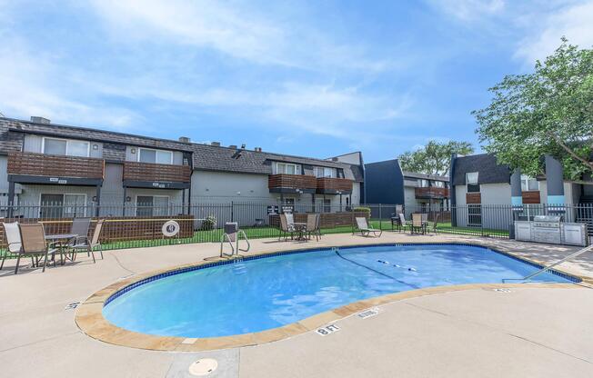 A clear blue swimming pool surrounded by lounge chairs and a grassy area. In the background, there are apartment buildings with balconies. The sky is bright and blue with a few clouds. A fence surrounds the pool area, providing a cozy atmosphere.