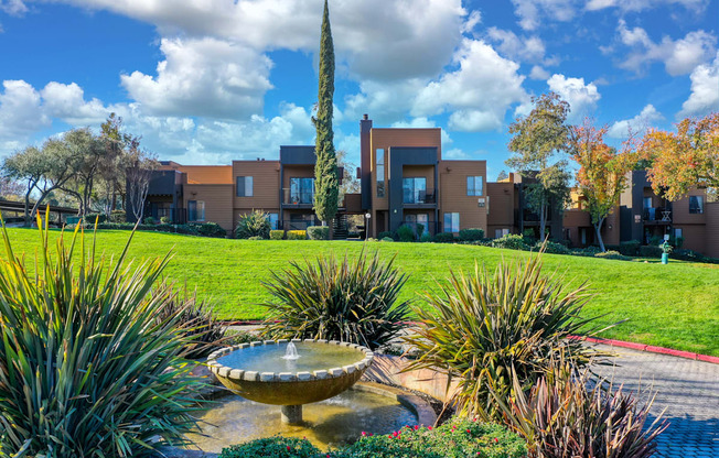 a fountain in the middle of a grassy area with apartment buildings in the background
