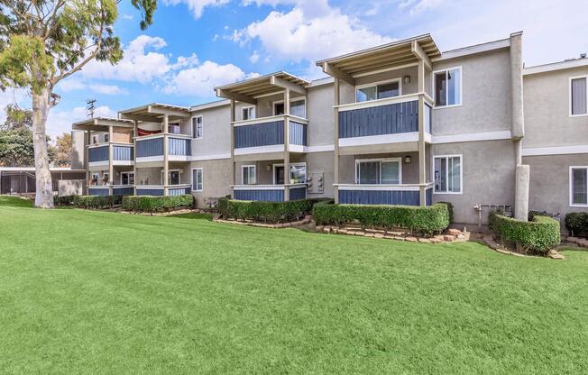 A row of modern apartment buildings with balconies, situated on a well-maintained green lawn. The sky is partly cloudy, and there are trees nearby, adding to the pleasant outdoor setting.