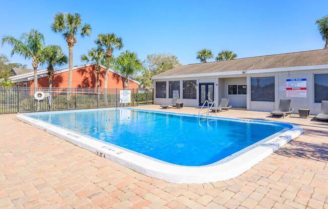 A clear blue swimming pool surrounded by a brick patio, with lounge chairs positioned nearby. Palm trees are visible in the background, and a building with a gray roof and multiple windows is adjacent to the pool area. The sky is bright and sunny.