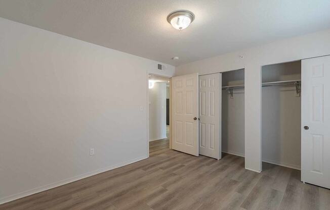 A room with a white wall and a wooden floor with a door on the left and two closets on the right.