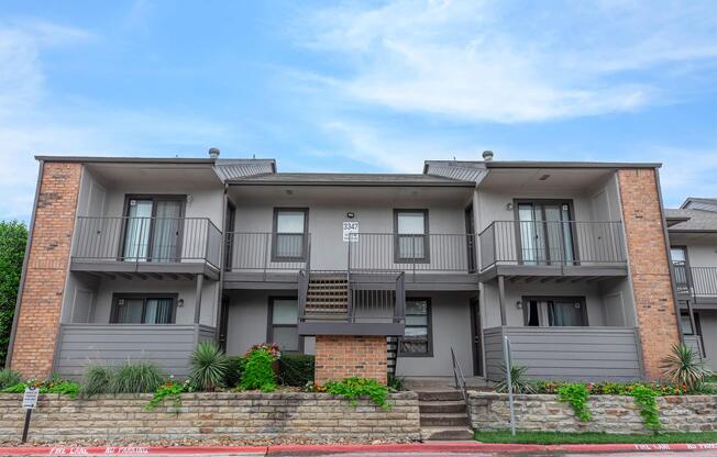 A two-story apartment building featuring a brick and gray exterior, with a central staircase leading to the upper units. Each unit has a balcony, and there are landscaped plants in front of the building. The sky above is clear and blue.