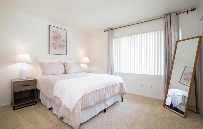 A cozy bedroom featuring a bed with light-colored linens and decorative pillows, a nightstand with a lamp, sheer window curtains, and a large mirror. The walls are painted in soft tones, and a piece of floral artwork decorates one wall, creating a serene and inviting atmosphere.