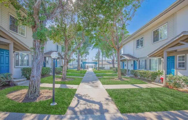 A walkway between two multi-unit residential buildings, featuring green lawns, well-maintained shrubs, and trees. The buildings have light-colored exteriors and blue doors, with a pathway leading towards a pool area in the background, surrounded by additional greenery and blue sky.