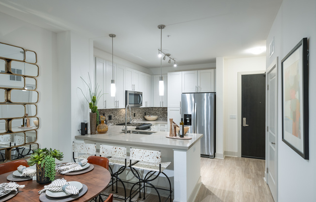 Open-concept kitchen and dining area with white walls, light wood-style floors, a four-person dining table, three barstools at the kitchen peninsula, white cabinets, and stainless steel appliances.