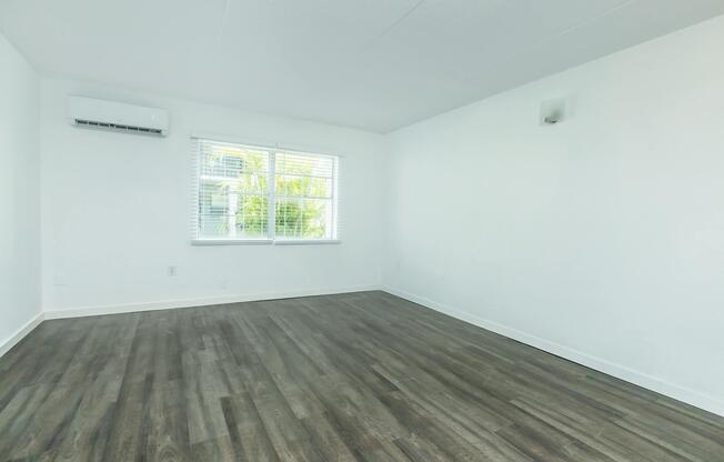 Empty room featuring light-colored walls and a single window with blinds. The floor is covered in dark wood laminate. An air conditioning unit is visible on the wall, and thereâs a light fixture mounted near the ceiling. The overall atmosphere is bright and minimalistic.