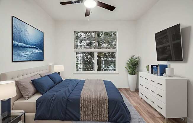 A bedroom with a bed, a ceiling fan, a window with a view of trees, a nightstand with a lamp, and a painting on the wall.
