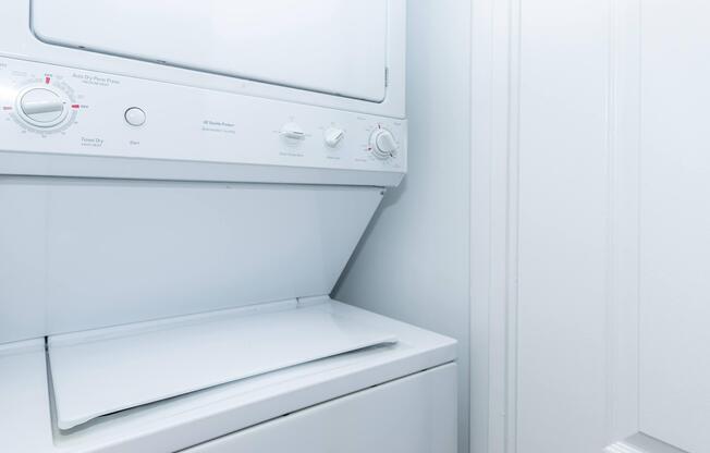A stacked washer and dryer unit in a laundry room, featuring a white exterior and control knobs for various settings. The walls are plain and light-colored, creating a clean and minimalist appearance.