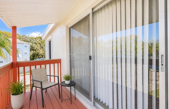 A small balcony or patio featuring a chair and side table, with potted plants. There are vertical blinds covering a large window, and a view of nearby buildings and trees in the background under a partly cloudy sky. The flooring is wooden with a reddish hue.