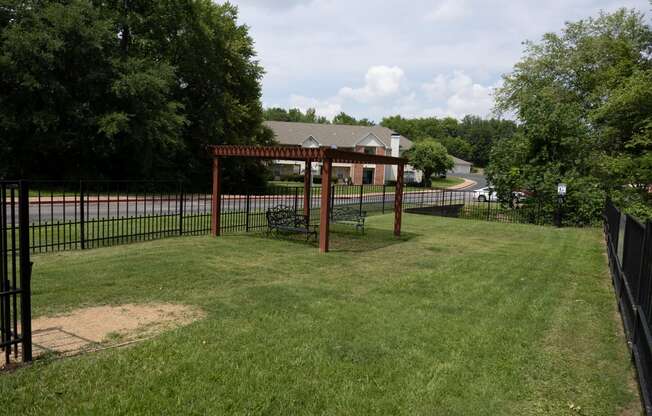 View from in gated dog park with pergola and seating area with wooded area and building in background