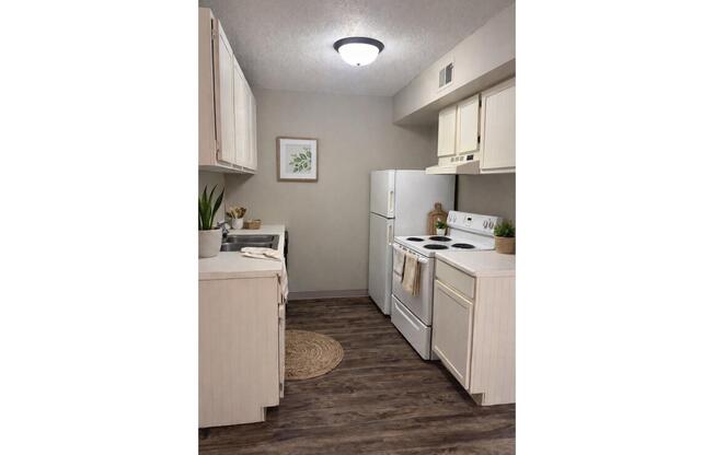 Modern kitchen with light-colored cabinets, white appliances, and wooden flooring. There is a small round rug on the floor, a plant on the counter, and framed artwork on the wall. The overall design is bright and inviting, showcasing a functional and stylish cooking space.