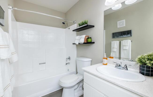 A clean and modern bathroom featuring a white bathtub with shower, a toilet, and a sink with a white countertop. There are neatly arranged towels and decor items on shelves, along with a small plant. The walls are painted a soft beige, enhancing the bright and tidy atmosphere.