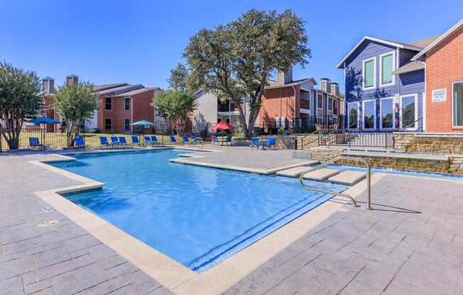 A sunny outdoor swimming pool area featuring a clear blue pool with lounge chairs, surrounded by greenery. In the background, there are several residential buildings with colorful exteriors, and shaded seating options under red umbrellas. The scene conveys a relaxing and inviting atmosphere.