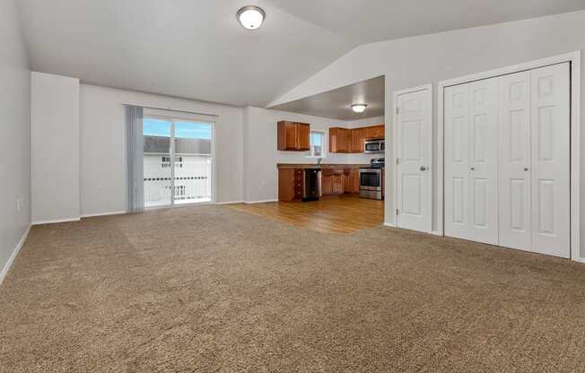 A spacious living room with a carpeted floor and a kitchen area in the background.
