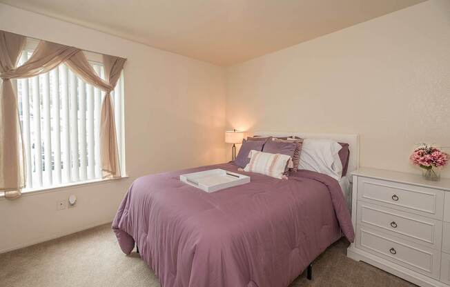 Model primary bedroom with a purple bedspread and a white nightstand at Pheasant Pointe Apartments, California, 95833