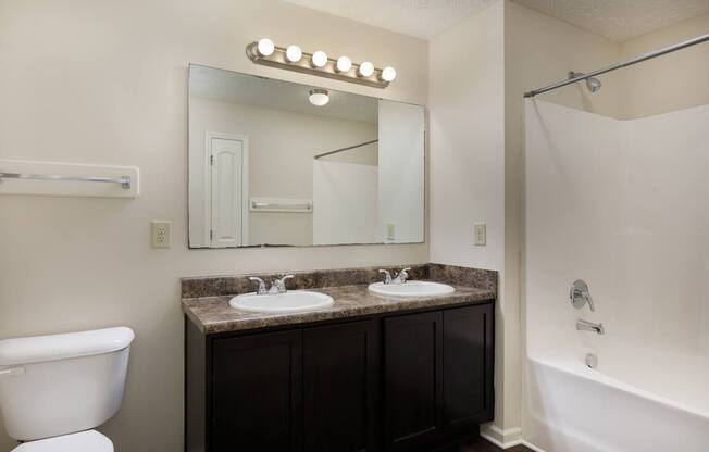 A bathroom with a white toilet, a sink with a marble countertop, a mirror with lights above it, and a bathtub with a shower curtain.